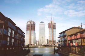 W11	Canary Wharf tower with the two new towers under construction, framed between some buildings.